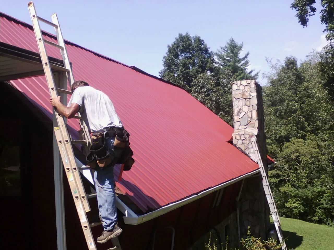 Roofer on a ladder beside a red metal roof and stone chimney — Soffit & Fascia Repair by Roofing Brothers in Manning